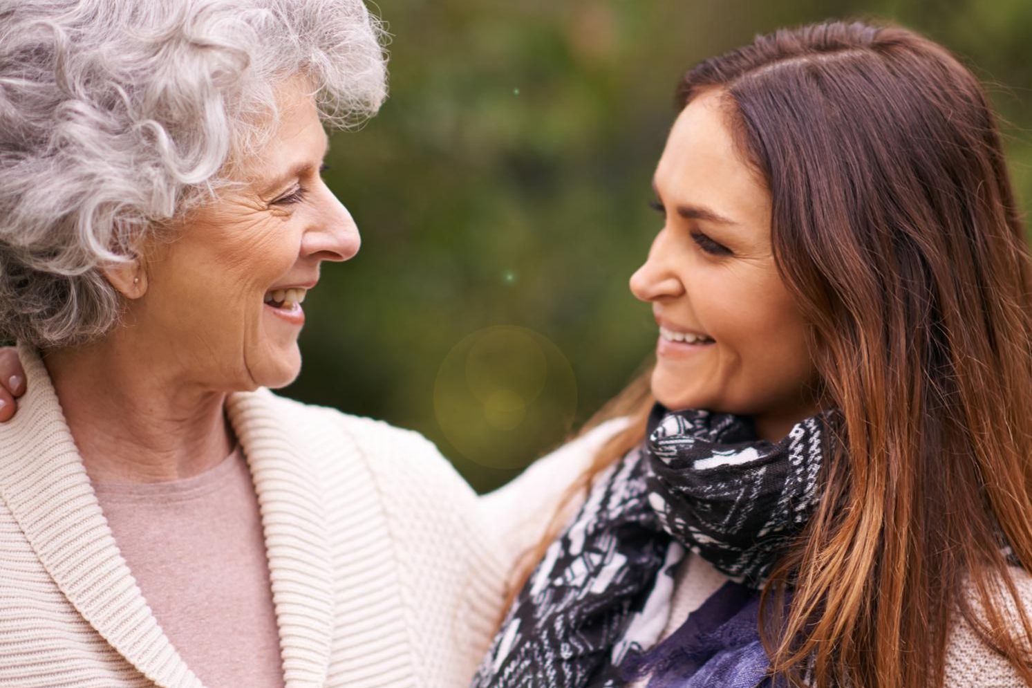 Woman with gray hair and daughter with long, brown hair looking at each other while smiling.