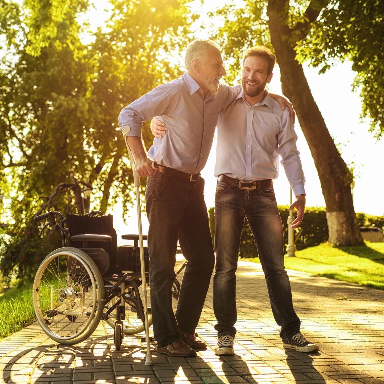 Older man is standing with his arm around a younger man in a park with green trees.